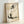 May include: Framed artwork leaning against white wall showing watercolor-style illustration of a plump, sitting cat in black and gray ink wash technique. Features Japanese calligraphy characters at top in bold black brushstrokes. Art has aged, textured paper appearance with subtle staining and speckles. Simple light wood frame surrounds the piece, casting soft shadow on wall.