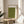 May include: Framed poster with olive green textured background featuring three small black bird silhouettes in flight and vertical Japanese calligraphy text on cream matting. Natural wood frame displayed on round wooden side table next to beige upholstered chair with white pillow. Soft natural lighting creates shadows on neutral wall with plant leaves visible in corner.