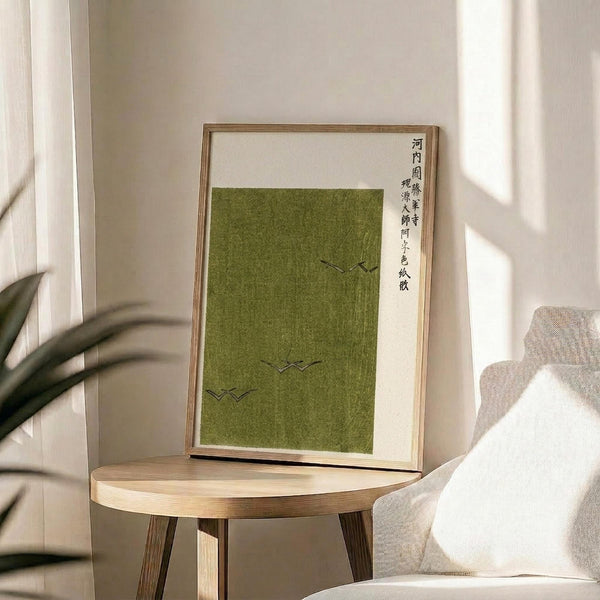 May include: Framed poster with olive green textured background featuring three small black bird silhouettes in flight and vertical Japanese calligraphy text on cream matting. Natural wood frame displayed on round wooden side table next to beige upholstered chair with white pillow. Soft natural lighting creates shadows on neutral wall with plant leaves visible in corner.