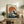 May include: Framed Japanese-style wave artwork with large orange-red sun behind dramatic blue and white crashing waves, displayed on rustic wooden sideboard alongside potted ferns, ceramic vases, and books in neutral-toned living room setting with beige chair and cream walls