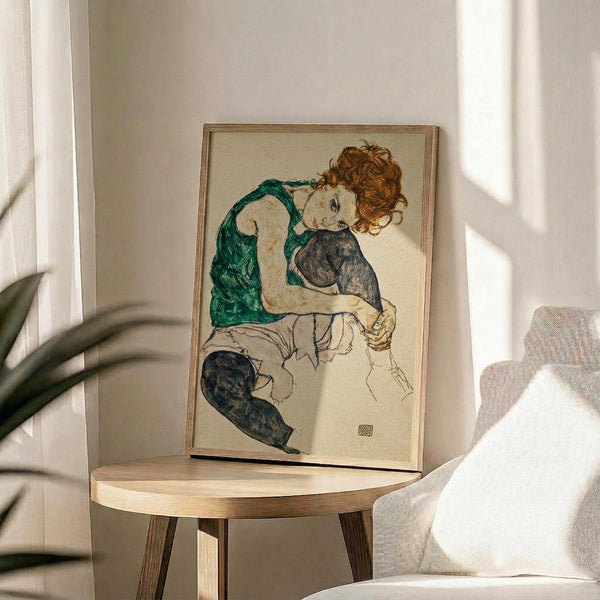 May include: Framed reproduction of an expressionist drawing showing a seated woman with curly reddish hair wearing a green top and dark stockings, displayed on a round wooden side table next to a beige upholstered chair with plant leaves visible in soft natural lighting