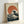 May include: Framed poster print leaning against white wall showing stylized ocean wave with white foam cresting toward large orange-red sun in background. Dark blue-green water dominates lower portion with dramatic wave formations. Japanese characters visible in upper right corner. Natural wood frame surrounds artwork on textured cream background. Print displays classic Japanese woodblock aesthetic with bold colors and flowing lines.