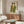 May include: Framed artwork showing two figures in green and yellow robes displayed above a wooden sideboard in a modern living room with cream walls, white curved chair, oval glass coffee table, and warm natural lighting casting shadows across the space.