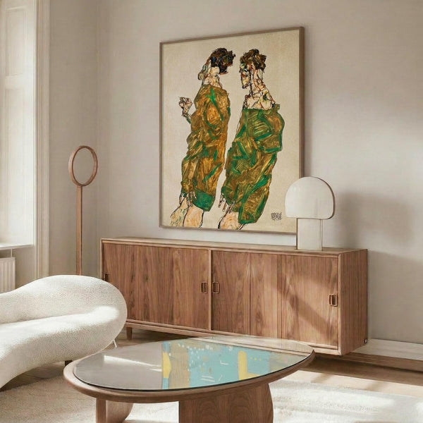 May include: Framed artwork showing two figures in green and yellow robes displayed above a wooden sideboard in a modern living room with cream walls, white curved chair, oval glass coffee table, and warm natural lighting casting shadows across the space.