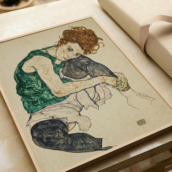 Drawing shows a seated woman with curly auburn hair wearing a green draped garment over white underclothes and dark stockings. The figure is rendered in Schiele's distinctive expressionist style with bold outlines and muted colors on cream paper. The artwork appears in an open art book or portfolio with visible binding and pages, photographed on a light wooden surface with soft natural lighting.