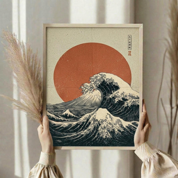 May include: Person holding framed Japanese-style artwork featuring a large wave with white foam crests against an orange-red sun on cream background. The print shows Hokusai-inspired woodblock aesthetic with dark blue-green water and traditional Asian characters in corner. Light wood frame, neutral beige clothing, dried pampas grass visible, soft natural lighting by window with white curtains.