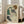May include: Framed reproduction of Schiele's expressionist drawing showing a seated woman with curly auburn hair wearing a green sleeveless top and dark stockings, holding a small dark object. The artwork features muted earth tones on cream background with visible aging spots. Large wooden-framed print leans against white wall next to beige upholstered chair in bright, naturally-lit room with hardwood floors.