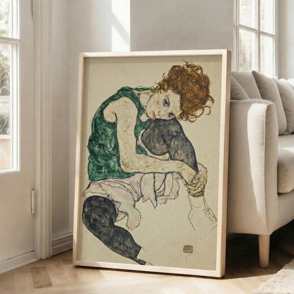 May include: Framed reproduction of Schiele's expressionist drawing showing a seated woman with curly auburn hair wearing a green sleeveless top and dark stockings, holding a small dark object. The artwork features muted earth tones on cream background with visible aging spots. Large wooden-framed print leans against white wall next to beige upholstered chair in bright, naturally-lit room with hardwood floors.