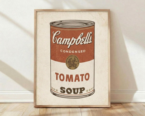 May include: Framed poster print of Campbell's condensed tomato soup can in Andy Warhol pop art style, featuring red and white label with vintage typography, displayed in thin wooden frame leaning against white wall on light wood floor with natural shadows