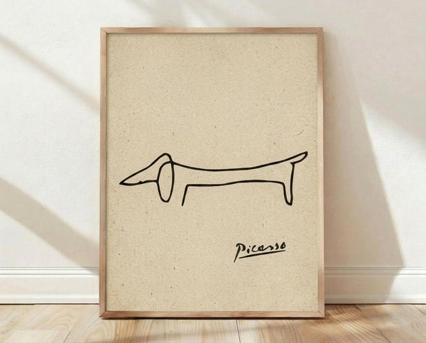 May include: Framed minimalist line drawing of a dachshund dog in black ink on beige textured paper background, displayed in light wood frame against white wall on wooden floor with natural lighting and shadows