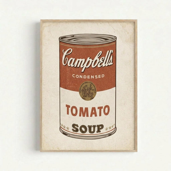 A framed print of a Campbell's condensed tomato soup can in the iconic pop art style. The artwork features a red and white soup can with "Campbell's" in white script lettering on the red upper portion and "TOMATO SOUP" in red text on the white lower portion. The print has a vintage, slightly weathered appearance and is displayed in a simple light wood frame against a white wall.