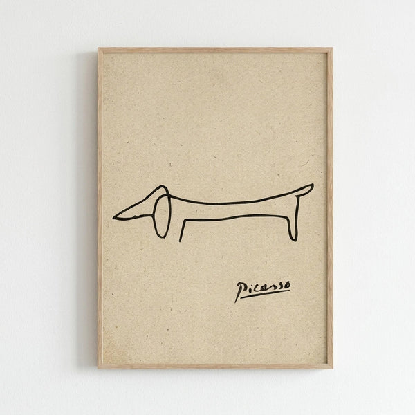 May include: Framed minimalist line drawing of a dachshund dog in black ink on cream textured paper background, displayed on white wall. Simple wooden frame surrounds the elongated dog illustration drawn with continuous flowing lines, with "Picasso" signature below the artwork.