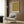 May include: Ornate golden-yellow botanical pattern poster with intricate floral and vine motifs in white frame, displayed on wood-paneled wall above cream upholstered armchair with geometric throw pillow, brass floor lamp, and dark round side table with open book in modern neutral-toned room