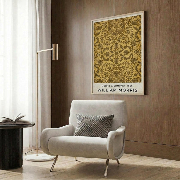 May include: Ornate golden-yellow botanical pattern poster with intricate floral and vine motifs in white frame, displayed on wood-paneled wall above cream upholstered armchair with geometric throw pillow, brass floor lamp, and dark round side table with open book in modern neutral-toned room