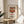May include: Framed poster print of Campbell's condensed tomato soup can in Andy Warhol's iconic pop art style, displayed on a light wood round side table next to a beige upholstered chair. The artwork features a red and white soup can with black text against a cream background in a thin wooden frame. Soft natural lighting creates shadows across the neutral-toned room with white walls and plant leaves visible.
