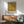 May include: Framed poster with ornate golden botanical pattern featuring symmetrical floral and leaf motifs in William Morris style, displayed above wooden sideboard with white ceramic lamp. Mid-century modern green upholstered chair with wooden arms sits nearby. Warm sunlight creates shadows on white wall in contemporary living room setting.
