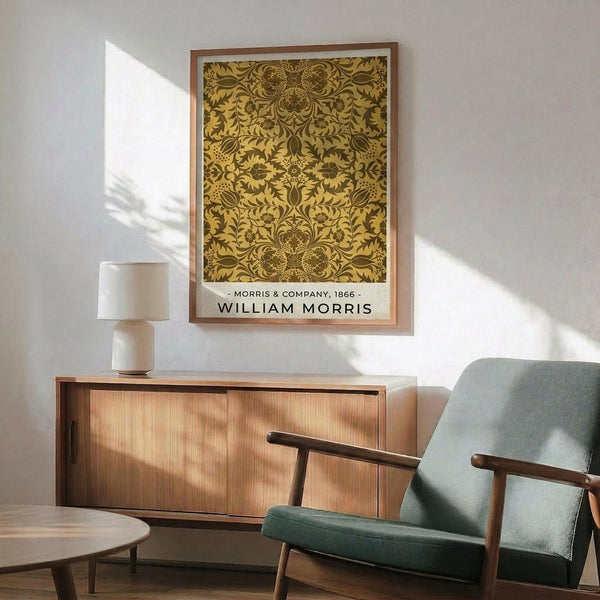 May include: Framed poster with ornate golden botanical pattern featuring symmetrical floral and leaf motifs in William Morris style, displayed above wooden sideboard with white ceramic lamp. Mid-century modern green upholstered chair with wooden arms sits nearby. Warm sunlight creates shadows on white wall in contemporary living room setting.