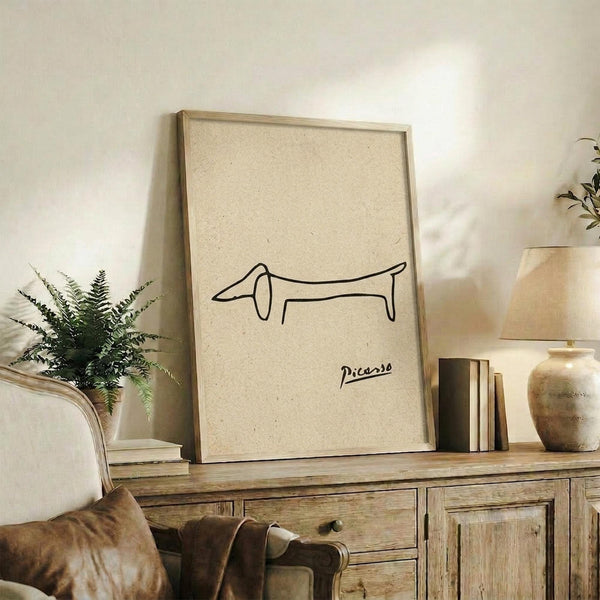 May include: Minimalist line drawing of a dachshund dog in black ink on cream textured paper, attributed to Picasso, displayed in light wood frame on rustic wooden sideboard alongside fern plant, ceramic vases, and stacked books in neutral-toned living room setting