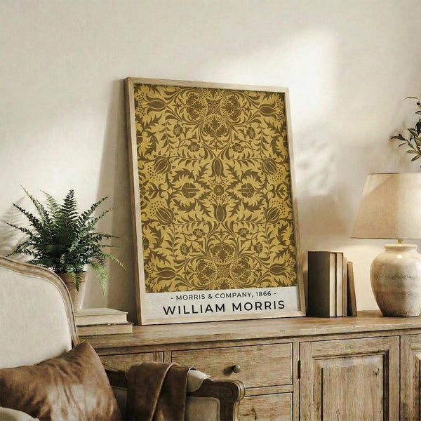 May include: Framed vintage botanical pattern poster with intricate golden yellow and brown floral damask design displayed on rustic wooden sideboard alongside fern plant, ceramic vase, books, and cream upholstered armchair in warm, naturally lit living room setting.