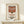 May include: Framed poster print of Campbell's condensed tomato soup can in red and white colors, displayed on light wood shelf against cream wall. Small white wrapped gift box with greenery sprig sits beside the natural wood frame. Vintage-style artwork shows classic soup can design with red top section, white bottom, and black lettering.
