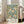 May include: Framed botanical art print leaning against beige armchair in bright living room with white walls and large windows. Print features vintage floral pattern with pink chrysanthemums, blue birds, and green foliage on cream background. Text below reads "Morris & Company, 1866 - William Morris" in black letters on white border.