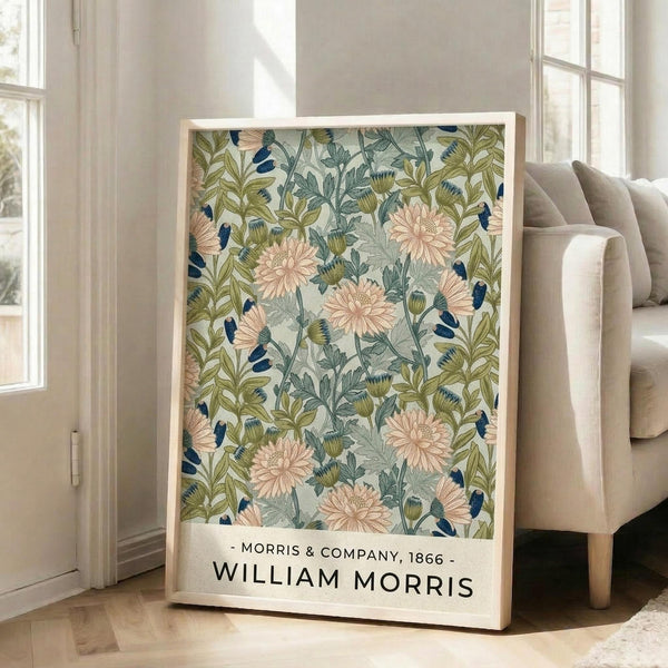 May include: Framed botanical art print leaning against beige armchair in bright living room with white walls and large windows. Print features vintage floral pattern with pink chrysanthemums, blue birds, and green foliage on cream background. Text below reads "Morris & Company, 1866 - William Morris" in black letters on white border.