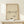 May include: Framed poster with minimalist line drawing of a dachshund dog and Picasso signature on beige textured paper background, displayed on light wood surface next to small white wrapped gift with evergreen sprig, against cream wall in natural lighting