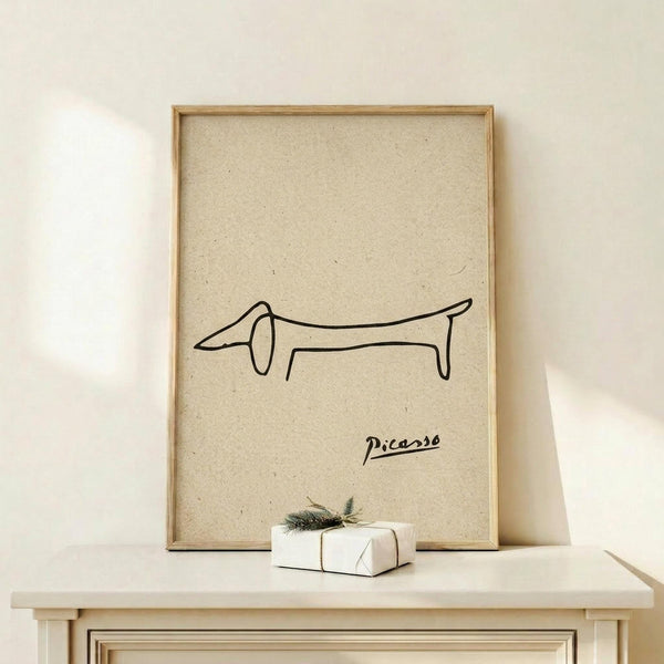 May include: Framed poster with minimalist line drawing of a dachshund dog and Picasso signature on beige textured paper background, displayed on light wood surface next to small white wrapped gift with evergreen sprig, against cream wall in natural lighting