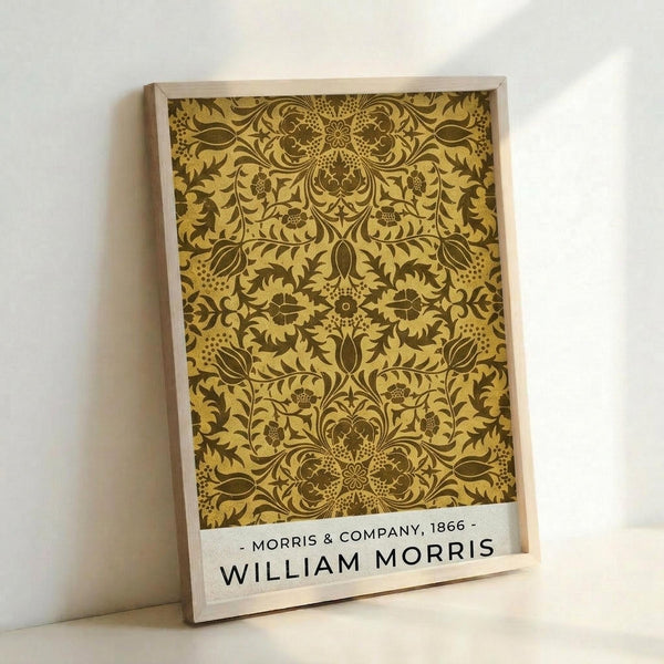 Ornate Victorian-style pattern poster in warm golden yellow and brown tones featuring intricate floral and botanical motifs with symmetrical damask design. The artwork displays detailed leaves, flowers, and decorative elements in a repeating pattern. Framed in light natural wood and leaning against a white wall, showing the full poster with white text attribution at bottom.