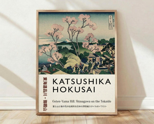 May include: Framed Japanese woodblock print reproduction featuring cherry blossom trees with pink blooms, rolling green hills, traditional buildings, and Mount Fuji in the distance. The artwork displays classic ukiyo-e style with blue-green sky and detailed landscape. White text below identifies the artist and location. Wooden frame sits on light wood floor against white wall with soft shadows.