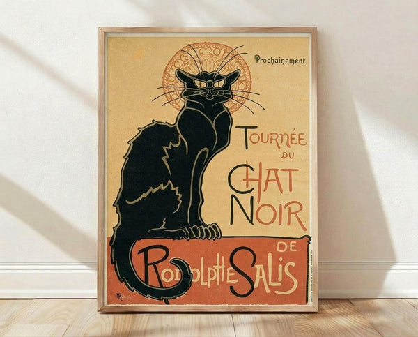 May include: Framed vintage French poster print featuring a black cat with arched back and flowing tail, sitting against cream background with ornate circular design behind its head. Red and orange text reads "Tournée du Chat Noir" and "Rodolphe Salis" with "Prochainement" at top. Art Nouveau style illustration in wooden frame, leaning against white wall on light wooden floor.