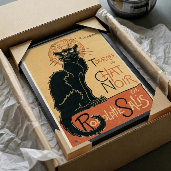 May include: Vintage French poster reproduction featuring iconic black cat silhouette with yellow eyes and ornate decorative halo, displayed in black frame within cardboard shipping box surrounded by white tissue paper and wooden dowels for protection. Art nouveau style artwork in warm yellow and orange tones with elegant French typography.