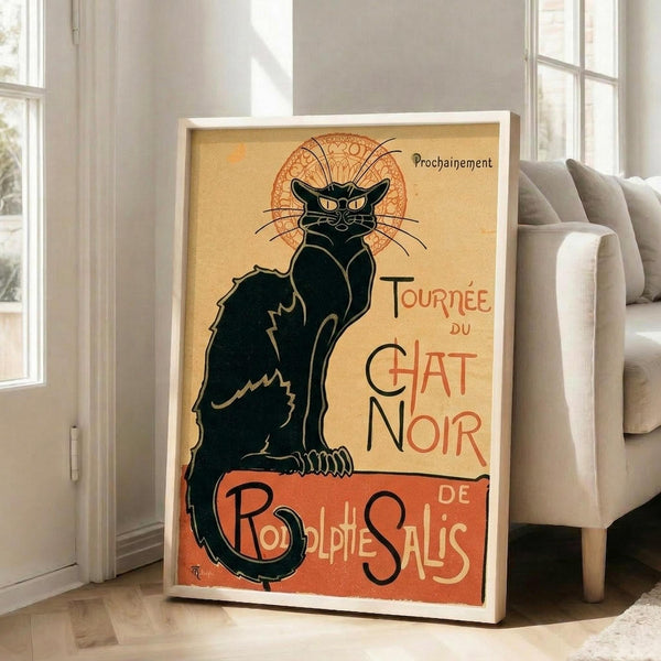 May include: Vintage-style poster print in white frame showing iconic black cat with yellow eyes sitting upright. Features Art Nouveau design with ornate circular backdrop and French text "Tournée du Chat Noir" and "Rodolphe Salis" in red lettering on cream and orange background. Frame leans against white wall next to beige upholstered chair in bright room with tall windows.