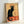 May include: Framed vintage French Art Nouveau poster featuring a black cat with arched back and yellow eyes against cream background with orange decorative elements. The stylized illustration shows bold red text reading "Tournée du Chat Noir" and "Rodolphe Salis" with ornate circular design behind the cat. Light wood frame sits propped against white wall, casting subtle shadows.