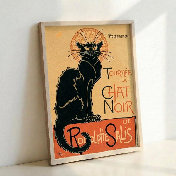 May include: Framed vintage French Art Nouveau poster featuring a black cat with arched back and yellow eyes against cream background with orange decorative elements. The stylized illustration shows bold red text reading "Tournée du Chat Noir" and "Rodolphe Salis" with ornate circular design behind the cat. Light wood frame sits propped against white wall, casting subtle shadows.