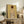 May include: Framed art print of a figure with reddish hair viewed from behind, rendered in earth tones on golden yellow background, displayed on rustic wooden console alongside potted fern, ceramic vessels, and stacked books in neutral-toned living room setting.