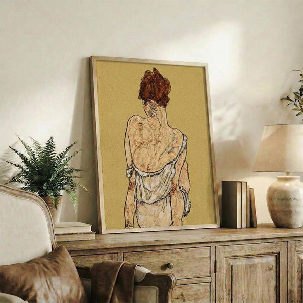May include: Framed art print of a figure with reddish hair viewed from behind, rendered in earth tones on golden yellow background, displayed on rustic wooden console alongside potted fern, ceramic vessels, and stacked books in neutral-toned living room setting.