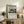 May include: Framed Japanese woodblock print featuring cherry blossom trees in pink bloom overlooking a blue landscape with Mount Fuji in the distance, displayed on a rustic wooden sideboard alongside potted fern, ceramic vase, and stacked books in a warm, naturally-lit living room setting with beige upholstered furniture.