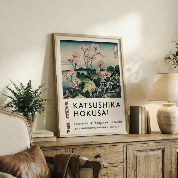 May include: Framed Japanese woodblock print featuring cherry blossom trees in pink bloom overlooking a blue landscape with Mount Fuji in the distance, displayed on a rustic wooden sideboard alongside potted fern, ceramic vase, and stacked books in a warm, naturally-lit living room setting with beige upholstered furniture.