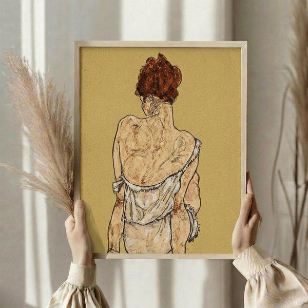 May include: Person in cream outfit holding framed art print showing seated figure from behind with upswept auburn hair against golden yellow background, displayed in light wooden frame. Dried wheat stems visible in left hand. Soft natural lighting through white curtains creates warm, minimalist interior setting.