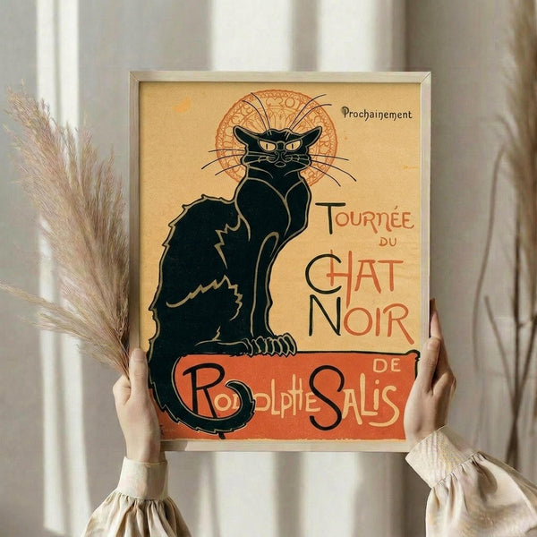 May include: Hands holding a framed vintage French poster featuring a black cat with yellow eyes sitting upright against an ornate orange circular background. The art nouveau style print displays "Tournée du Chat Noir" and "Rodolphe Salis" text in decorative lettering on a cream background. A bundle of dried pampas grass stems appears to the left, with neutral curtains softly blurred in the background.