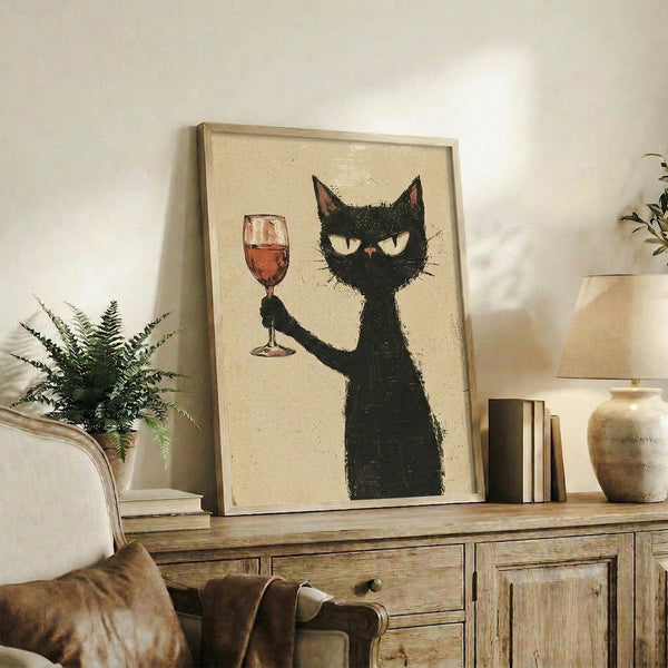 May include: Framed poster print featuring a stylized black cat with yellow eyes holding up a wine glass filled with red wine, displayed on a rustic wooden sideboard next to books, a fern plant, and ceramic vases in a cozy living room setting with neutral walls and brown leather furniture.