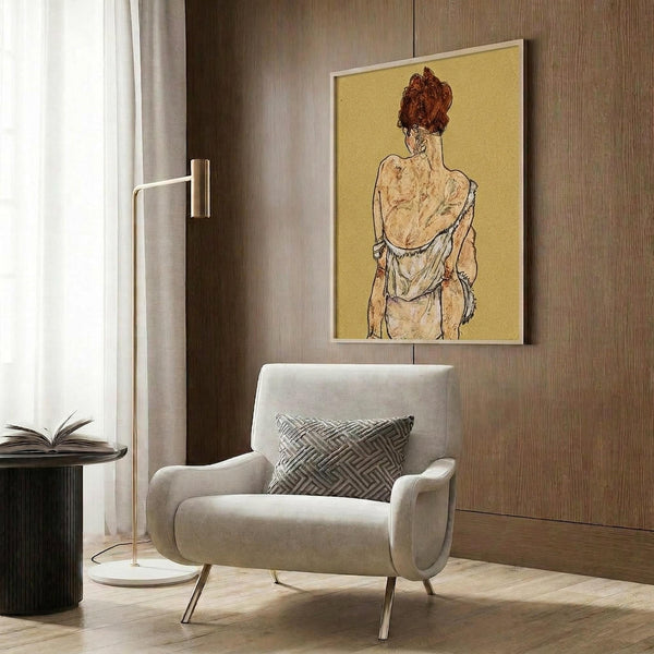 May include: Framed reproduction of Egon Schiele artwork showing figure with red hair from behind, draped in white fabric against yellow background. Modern living room setting with beige upholstered chair, geometric patterned pillow, gold floor lamp, dark round side table with open book, and wood paneled wall.
