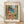 May include: Framed art print featuring a Mediterranean coastal scene with coral and peach colored buildings, green shutters, palm tree, blue water with small boats, viewed through an open window frame. The poster has "MATISSE" at top and "VIEW OF NICE" at bottom in terracotta lettering. Natural wood frame sits on light wooden floor against white wall with soft shadows.