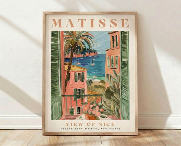 May include: Framed art print featuring a Mediterranean coastal scene with coral and peach colored buildings, green shutters, palm tree, blue water with small boats, viewed through an open window frame. The poster has "MATISSE" at top and "VIEW OF NICE" at bottom in terracotta lettering. Natural wood frame sits on light wooden floor against white wall with soft shadows.