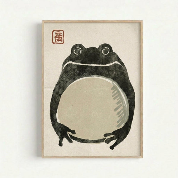 A stylized frog illustration in dark green ink with a large pale circular belly, rendered in traditional Japanese woodblock print style. The frog has prominent round eyes and sits upright with visible front legs. A red square seal with Japanese characters appears in the upper left corner. The artwork is displayed in a light wooden frame against a cream-colored background, mounted on a white wall.