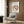 May include: Matisse "The Dance" poster in light wooden frame hanging on wood-paneled wall above beige upholstered armchair with geometric patterned pillow. Poster shows red dancing figures in circle formation on cream background. Modern brass floor lamp and dark round side table with open book visible. Neutral toned contemporary interior setting with natural lighting.