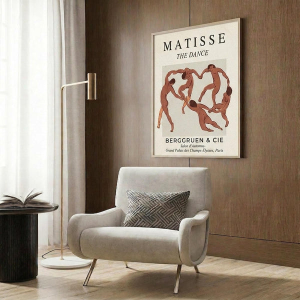 May include: Matisse "The Dance" poster in light wooden frame hanging on wood-paneled wall above beige upholstered armchair with geometric patterned pillow. Poster shows red dancing figures in circle formation on cream background. Modern brass floor lamp and dark round side table with open book visible. Neutral toned contemporary interior setting with natural lighting.