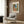 May include: A cream-colored upholstered armchair with geometric patterned pillow positioned against wood-paneled wall. Above hangs a framed Matisse poster showing a Mediterranean coastal scene with coral buildings, green shutters, palm trees and blue water. Modern brass floor lamp and dark round side table with open book complete the warm, contemporary reading nook setting.