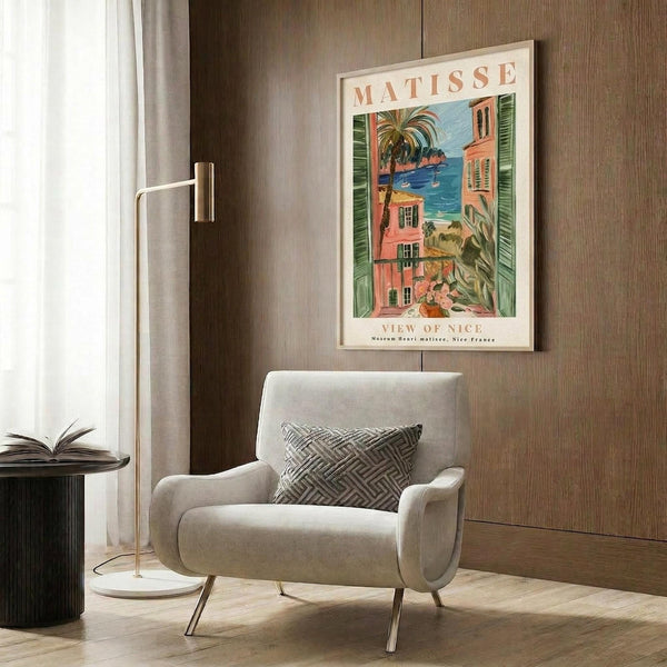 May include: A cream-colored upholstered armchair with geometric patterned pillow positioned against wood-paneled wall. Above hangs a framed Matisse poster showing a Mediterranean coastal scene with coral buildings, green shutters, palm trees and blue water. Modern brass floor lamp and dark round side table with open book complete the warm, contemporary reading nook setting.